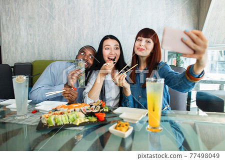 Selfie time, lunch and friendship concept. Pretty red haired girl taking a picture by the cell phone with her friends, Asian woman and African man, enjoying sushi food. Selfie time, lunch and friendship concept. Pretty red haired girl taking a picture by the cell phone with her friends, Asian woman and African man, enjoying sushi food. 77498049