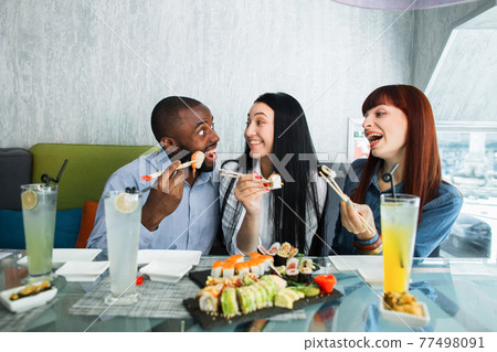 Happy three multiracial friends enjoying sushi at a Japanese restaurant. Pretty women and handsome man, eating sushi rolls and communicating, sitting at cafe. 77498091
