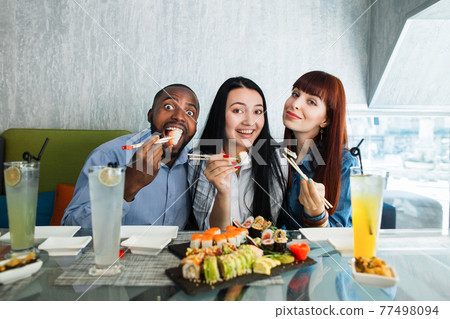 Sushi concept, seafood, asian meal. Three young multiethnic friends, Caucasian and Asian girls, African man, sitting in restaurant, spending funny time with plate of delicious sushi. Sushi concept, seafood, asian meal. Three young multiethnic friends, Caucasian and Asian girls, African man, sitting in restaurant, spending funny time with plate of delicious sushi. 77498094