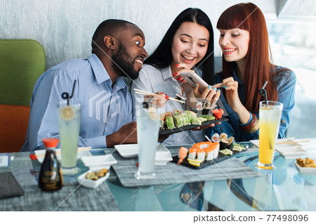 People, gadgets, food and blogging concept. Team of multicultural young people in cafe eating sushi rolls, having fun. Red haired Caucasian woman makes a photo of sushi on phone. People, gadgets, food and blogging concept. Team of multicultural young people in cafe eating sushi rolls, having fun. Red haired Caucasian woman makes a photo of sushi on phone. 77498096