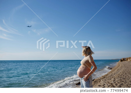 young pregnant girl by the sea looks at a flying plane 77499474