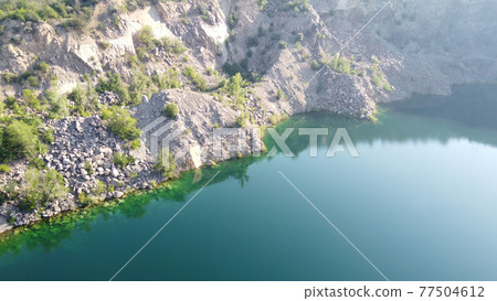 Rocky shore of the Radon Lake on a sunny summer morning. Aerial view of an old flooded granite quarry. A picturesque pond. Rocky shore of the Radon Lake on a sunny summer morning. Aerial view of an old flooded granite quarry. A picturesque pond. 77504612