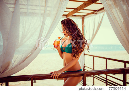 Portrait of sexy young woman in bikini with orange cocktail glass with straw and fruit posing at wooden bar of tent with white curtains. Portrait of sexy young woman in bikini with orange cocktail glass with straw and fruit posing at wooden bar of tent with white curtains. 77504634