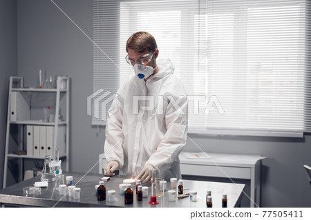 Young scientist in protective clothing and a respirator working in a chemistry laboratory, experimenting with heating substances, lighting up a match Young scientist in protective clothing and a respirator working in a chemistry laboratory, experimenting with heating substances, lighting up a match 77505411