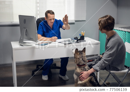 A male vet doctor in a blue uniform examining a husky dog in his clinic 77506139