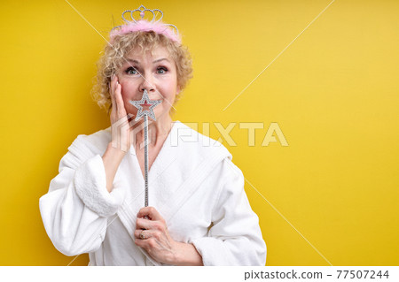 Beautiful elderly woman in bathrobe wearing crown on head, holding magica wand in hands, fairy 77507244