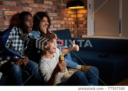 Emotional students watching sport match at home sitting on sofa. fans cheering for favourite national team, drinking beer Emotional students watching sport match at home sitting on sofa. fans cheering for favourite national team, drinking beer 77507380