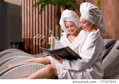 girlfriends in spa salon in towel and bathrobe looking at prices of procedures drinking champagne 77507843