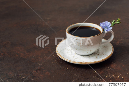Chicory drink in a cup and a blue chicory flower on a dark brown background. Coffee substitute. Space for text. Chicory drink in a cup and a blue chicory flower on a dark brown background. Coffee substitute. Space for text. 77507957