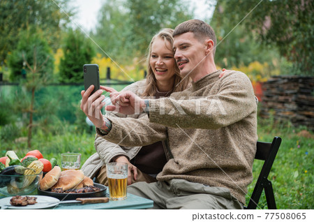 Husband and wife, young adults escaped from the city in a country house in nature to celebrate a family holiday date. Call by video on smartphone. Picnic with grill, beer and vegetables. Husband and wife, young adults escaped from the city in a country house in nature to celebrate a family holiday date. Call by video on smartphone. Picnic with grill, beer and vegetables. 77508065