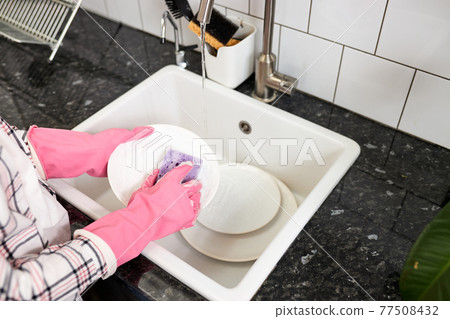Close up of female hands in pink protective rubber gloves washing white plate with purple cleaning sponge Close up of female hands in pink protective rubber gloves washing white plate with purple cleaning sponge 77508432