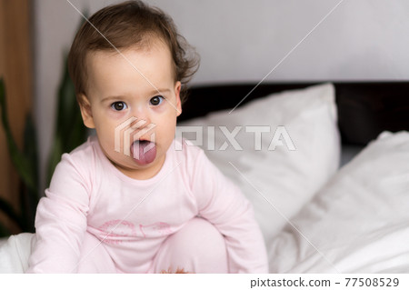 Authentic portrait cute caucasian little infant chubby baby girl or boy in pink sleepy upon waking looking at camera showing tongue in white bed. Child care, Childhood, Parenthood, lifestyle concept 77508529