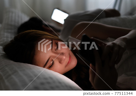Young couple conflict in bed. happy smiling woman turned her back to man, reading message on mobile phone, trying to peek at screen. Cheating and infidelity concept. Young couple conflict in bed. happy smiling woman turned her back to man, reading message on mobile phone, trying to peek at screen. Cheating and infidelity concept. 77511344