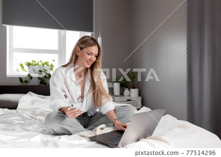 Smiling girl sits on the bed in bedroom and use phone and notebook 77514296