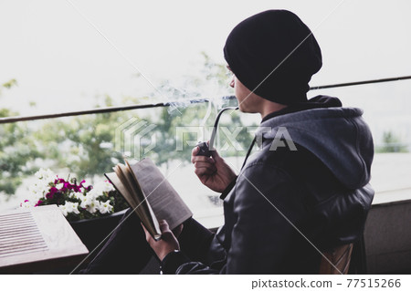 Back view on young black haired man witn piercing reeding a book and smoking tabacco pipe in outdoor cafe. 77515266