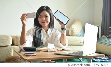 Photo of beautiful woman holding a credit card and white blank screen smartphone while sitting and relaxing in front of computer laptop that putting on wooden working desk over room as background. Photo of beautiful woman holding a credit card and white blank screen smartphone while sitting and relaxing in front of computer laptop that putting on wooden working desk over room as background. 77518026