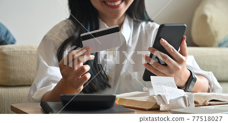 Cropped of smiling woman holding a credit card and smartphone while sitting in front of her calculator that putting on wooden table. 77518027