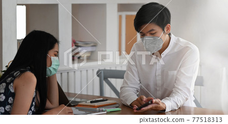 Photo of young couple that wearing a medical mask sitting and relaxing together at the wooden working desk over comfortable living room as background. 77518313