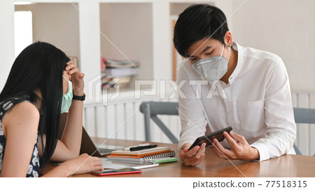 Photo of young couple that wearing a medical mask sitting and relaxing together at the wooden working desk over comfortable living room as background. Photo of young couple that wearing a medical mask sitting and relaxing together at the wooden working desk over comfortable living room as background. 77518315
