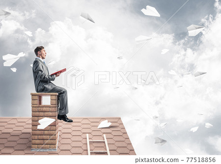 Man on brick roof reading book and paper planes flying in air Man on brick roof reading book and paper planes flying in air 77518856