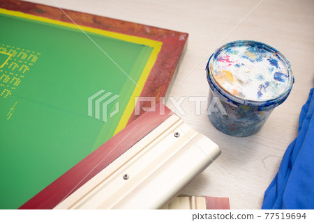 Serigraphy silk screen print process at clothes factory. Frame template stencil, squeegee and plastisol paint at table with cloth Serigraphy silk screen print process at clothes factory. Frame template stencil, squeegee and plastisol paint at table with cloth 77519694