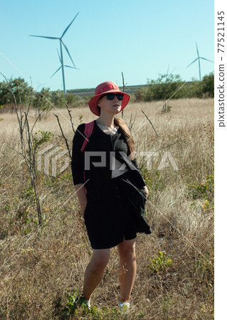 Woman in red hat in the field with wild grass blue sky wind turbines background. Stylish girl long brunette hair Woman in red hat in the field with wild grass blue sky wind turbines background. Stylish girl long brunette hair 77521145