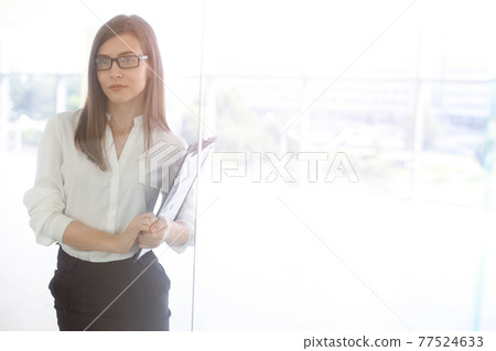 Beautiful female specialist with clipboard standing in sunny office and smiling charmingly. Business people concept 77524633