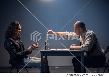 A detained white man with long hair receives irrefutable evidence of his guilt from a criminal investigator at the police station A detained white man with long hair receives irrefutable evidence of his guilt from a criminal investigator at the police station 77526855
