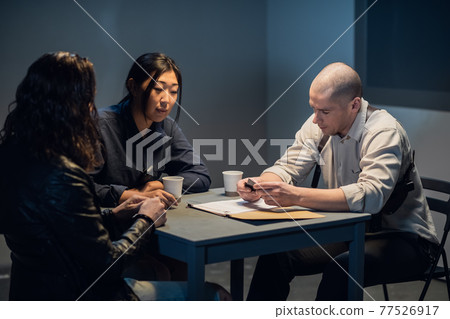 In the interrogation room, the investigator and the officer are talking to the criminal In the interrogation room, the investigator and the officer are talking to the criminal 77526917