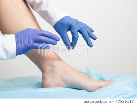 The doctor removes wax strips from the hair of the patient's legs on a white background. Leg hair removal concept, close-up The doctor removes wax strips from the hair of the patient's legs on a white background. Leg hair removal concept, close-up 77529117