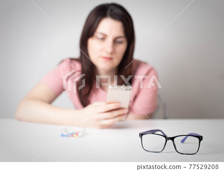 The girl looks at the smartphone screen and glasses from the view lie in the foreground. The harm of a smartphone to human vision The girl looks at the smartphone screen and glasses from the view lie in the foreground. The harm of a smartphone to human vision 77529208