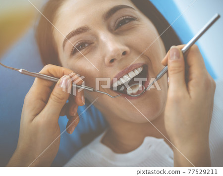Smiling brunette woman being examined by dentist at dental clinic. Hands of a doctor holding dental instruments near patient's mouth. Healthy teeth and medicine concept 77529211