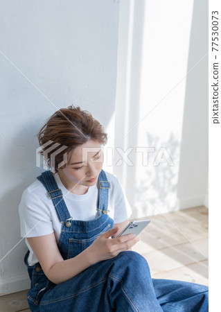 身著工裝褲的無性別 LGBT 男孩女孩,一位年輕女子坐在地板上摸著她的智能手機 身著工裝褲的無性別 LGBT 男孩女孩,一位年輕女子坐在地板上摸著她的智能手機 77530073