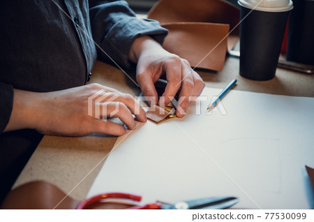 Tailor's hands make loops for the fastener of a women's bag made of genuine leather. Workbench in the workshop Tailor's hands make loops for the fastener of a women's bag made of genuine leather. Workbench in the workshop 77530099