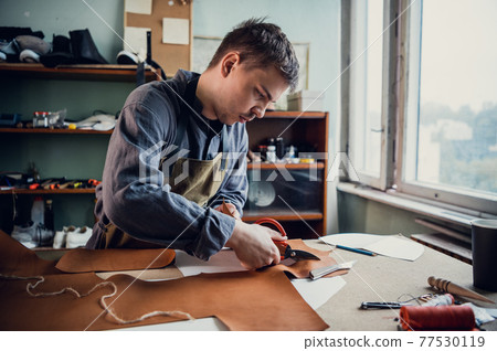 A talented young guy helps his father in the family business, cuts out blanks for shoes from leather A talented young guy helps his father in the family business, cuts out blanks for shoes from leather 77530119