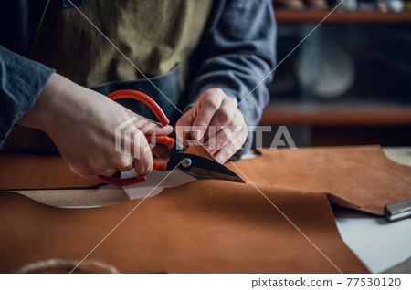 At the masterclass for the production of shoes, an experienced shoemaker cuts out leather with scissors At the masterclass for the production of shoes, an experienced shoemaker cuts out leather with scissors 77530120