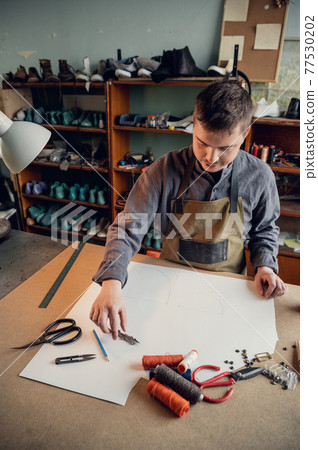 An experienced shoemaker in a shoe production workshop makes a pattern for a shoe from paper with different tools 77530202