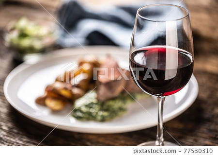 Red wine as a typical baverage to a red meat steak with roasted potatoes, spinach dip and fresh salad served on a white porcelain plate on the wooden desk. 77530310