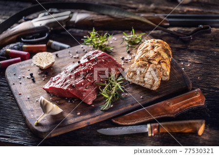 Nutrient rich raw deer venison prepared for a cooking process on a rustic wooden desk with roasted garlic, rosemary and huntig accesories like shot gun and ammunition 77530321