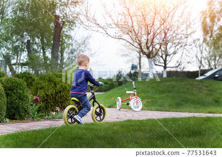 Back view little cute adorable caucasian toddler boy having fun riding exercise balance run bike push scooter in park or yard garden forest. Child first bike. Kids outdoor sport summer activities 77531643