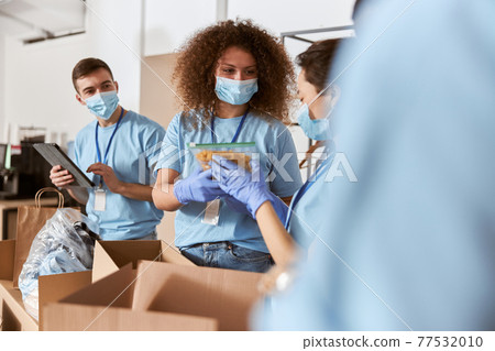 Busy volunteers wearing blue uniform, protective masks and gloves sorting donated pack of pasta and other food items while working in charitable foundation Busy volunteers wearing blue uniform, protective masks and gloves sorting donated pack of pasta and other food items while working in charitable foundation 77532010