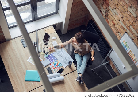 High angle view of female interior designer taking headphones, reading a blueprint while working on new project, sitting in modern office High angle view of female interior designer taking headphones, reading a blueprint while working on new project, sitting in modern office 77532362