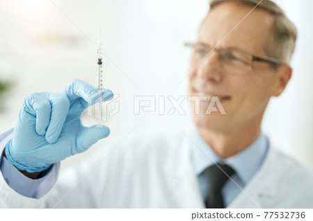 Cheerful male doctor holding syringe with vaccine Cheerful male doctor holding syringe with vaccine 77532736