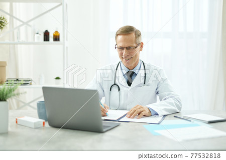 Cheerful doctor using laptop and filling in patient medical chart Cheerful doctor using laptop and filling in patient medical chart 77532818