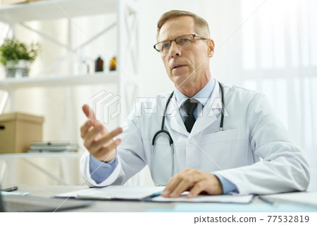 Handsome doctor sitting at the table in his office Handsome doctor sitting at the table in his office 77532819