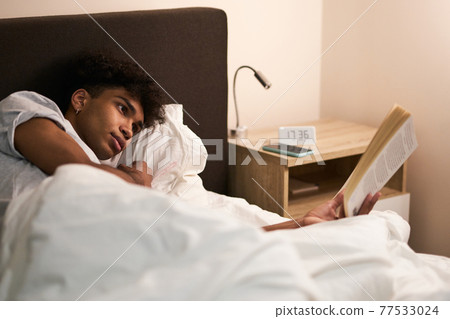 Young guy lying in the bed at home, resting and reading an interesting book before going to sleep 77533024