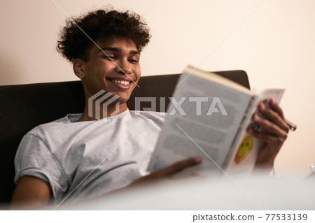Amazing story. Portrait of cheerful brunette guy lying in the bed at home, smiling while reading interesting book at late evening 77533139