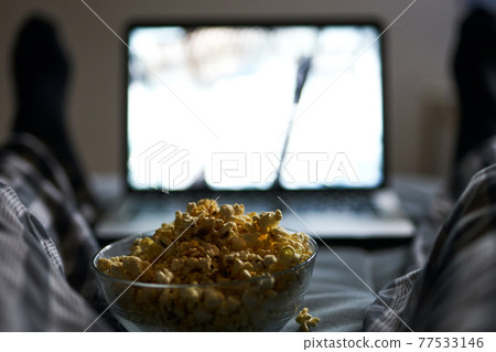 Its time to chill. Close up shot of popcorn in a bowl. Man in pajamas watching online movie using laptop, lying in the bed at home in the evening 77533146