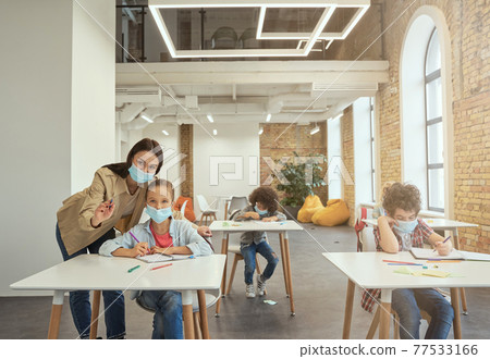 Attractive young female teacher wearing protective face mask helping little girl, explaining task. Kids studying in elementary school, sitting at the desk Attractive young female teacher wearing protective face mask helping little girl, explaining task. Kids studying in elementary school, sitting at the desk 77533166