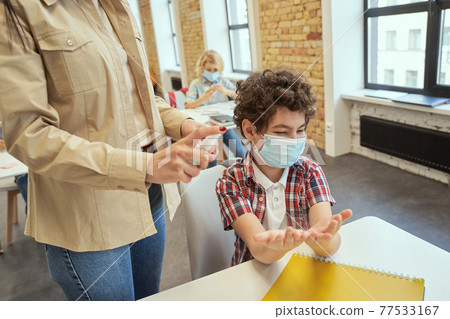 Little school boy wearing protective mask ready for cleaning his hands. Female teacher using an alcohol spray to disinfect student hands in classroom Little school boy wearing protective mask ready for cleaning his hands. Female teacher using an alcohol spray to disinfect student hands in classroom 77533167